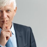 Business Lifestyle. Businessman standing isolated on gray showing secret gesture looking camera playful close-up