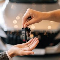 Close-up hands of unrecognizable dealer male giving new car key to customer man in business suit on blurred background of new auto in dealership office. Concept of choosing and buying car at showroom.
