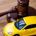 A close-up image of a yellow toy car and a wooden judge's gavel on a wooden table. Transport law, lemon law concept. A close-up image of a yellow toy car and a wooden judge's gavel on a wooden table. Transport law, lemon law concept.