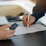 Closeup shot of customer signing contract and salesman holding car keys in a dealership center, unrecognizable man buying vehicle in auto showroom, filling documents, cropped