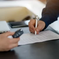Closeup shot of customer signing contract and salesman holding car keys in a dealership center, unrecognizable man buying vehicle in auto showroom, filling documents, cropped